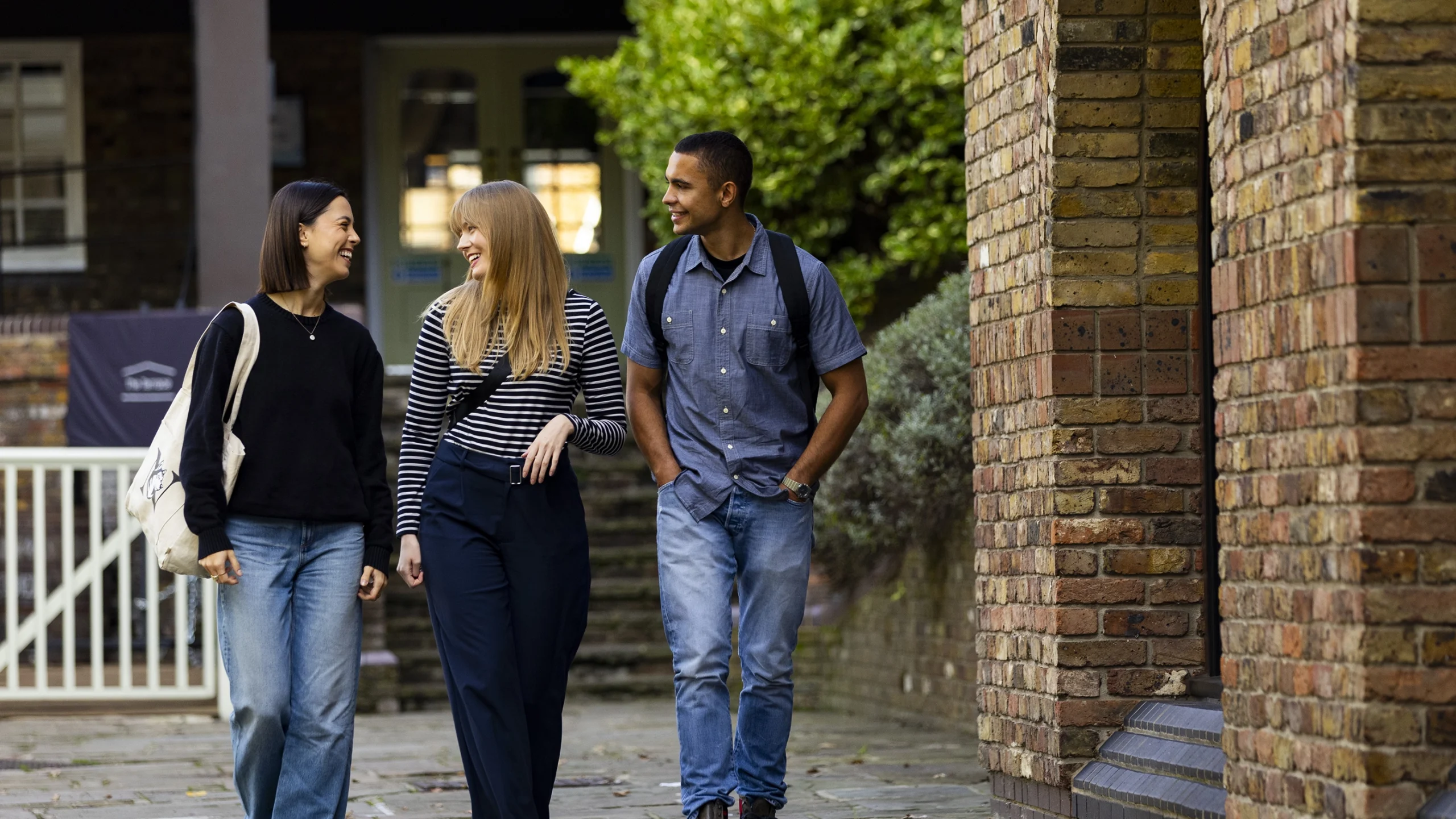 Three young commuter students, two women and one man, walk together outside along a brick building. Smiling and casually dressed, they appear to be having a friendly conversation with greenery visible in the background.