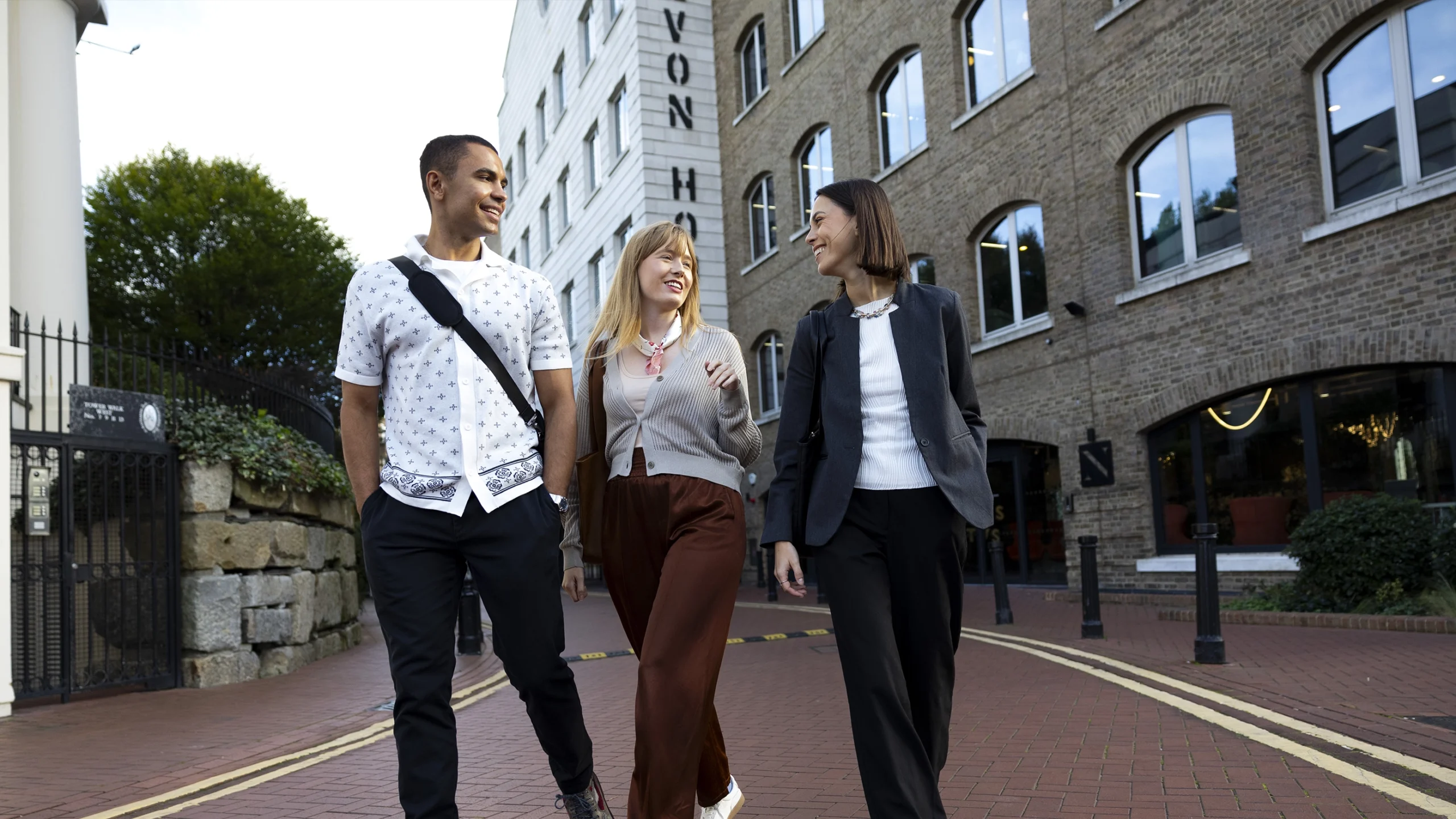 Three people, two women and one man, walk and chat together on a brick path by a modern building with large windows, appearing relaxed and happy as they discuss Master’s Tuition Fees.