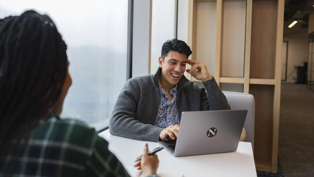 Two people sit at a table by a large window; one is smiling at a laptop, while the other faces away with a pen and notebook, suggesting a casual discussion about Master’s tuition fees.
