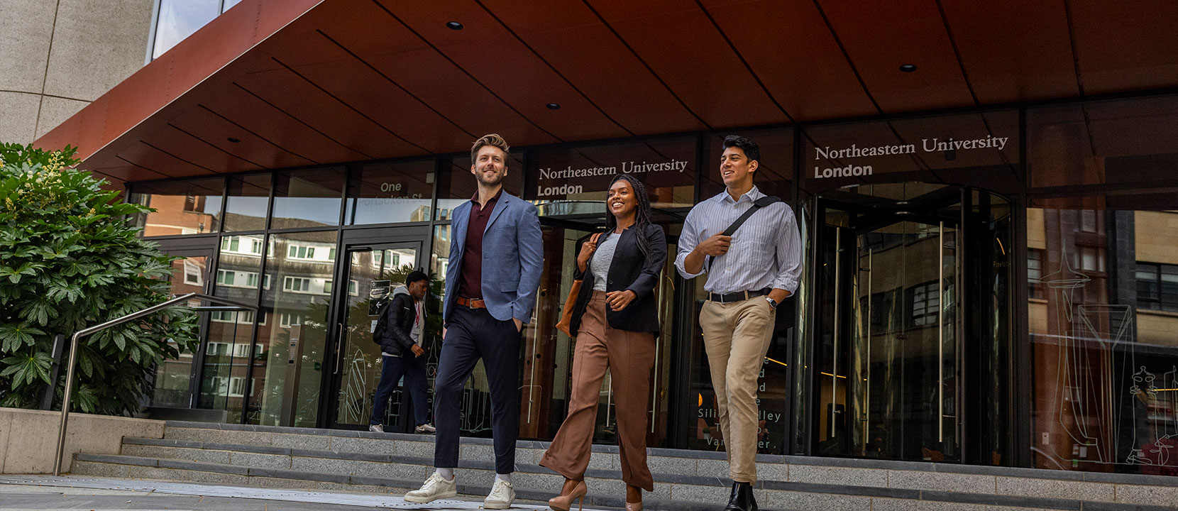 Three people in business attire walk and smile outside the entrance of Northeastern University London, with the university name above the glass doors—highlighting a welcoming environment for those pursuing masters-degrees.