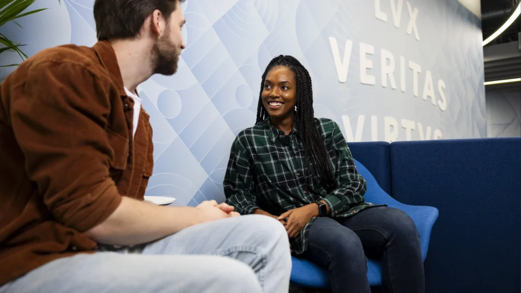 Two people sit and talk in a modern lounge with blue chairs and a patterned wall behind them featuring large white words, including "VERITAS." The woman smiles as they discuss Master’s tuition fees while the man listens.