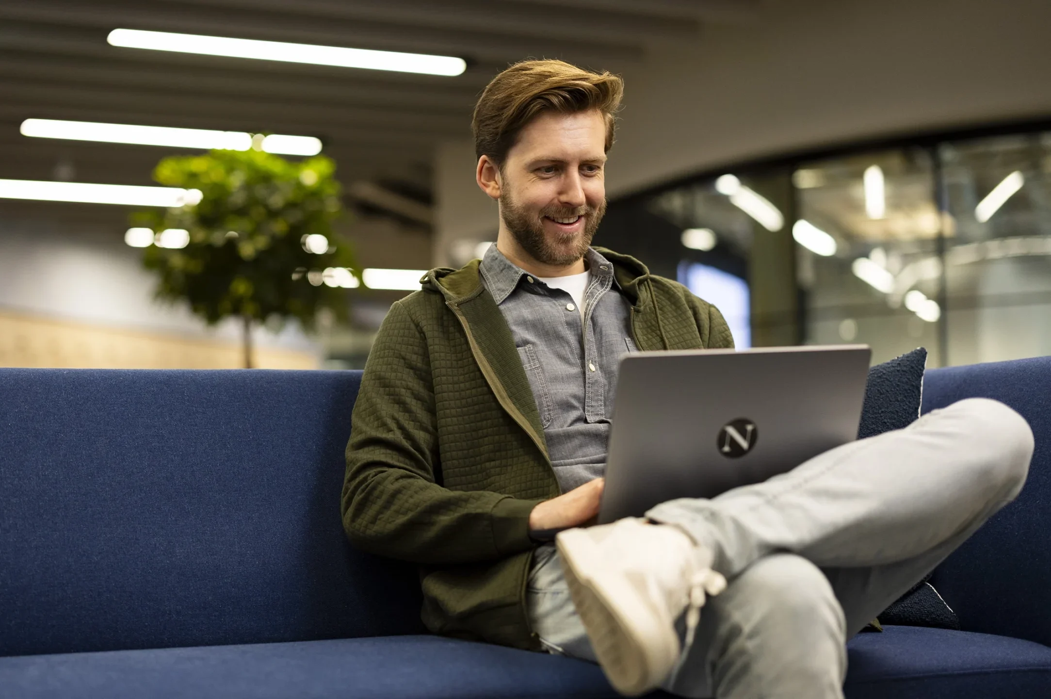 A man sits comfortably on a blue sofa, smiling as he reviews Master’s Tuition Fees on his laptop. Casually dressed in a hoodie and jeans, with his legs crossed, he relaxes in a modern, brightly lit office space.