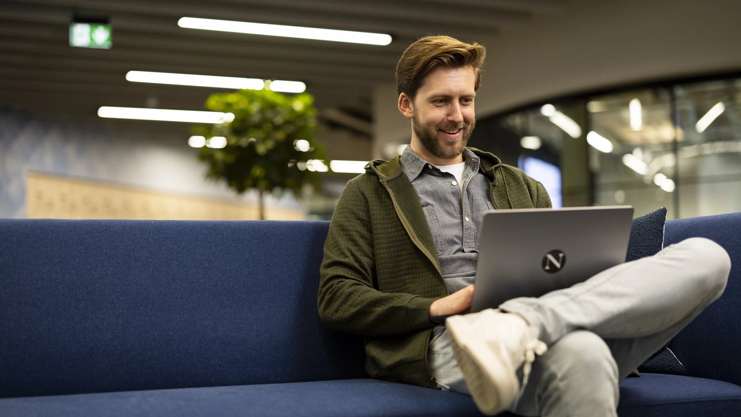 A man sits comfortably on a blue sofa, smiling as he reviews Master’s Tuition Fees on his laptop. Casually dressed in a hoodie and jeans, with his legs crossed, he relaxes in a modern, brightly lit office space.