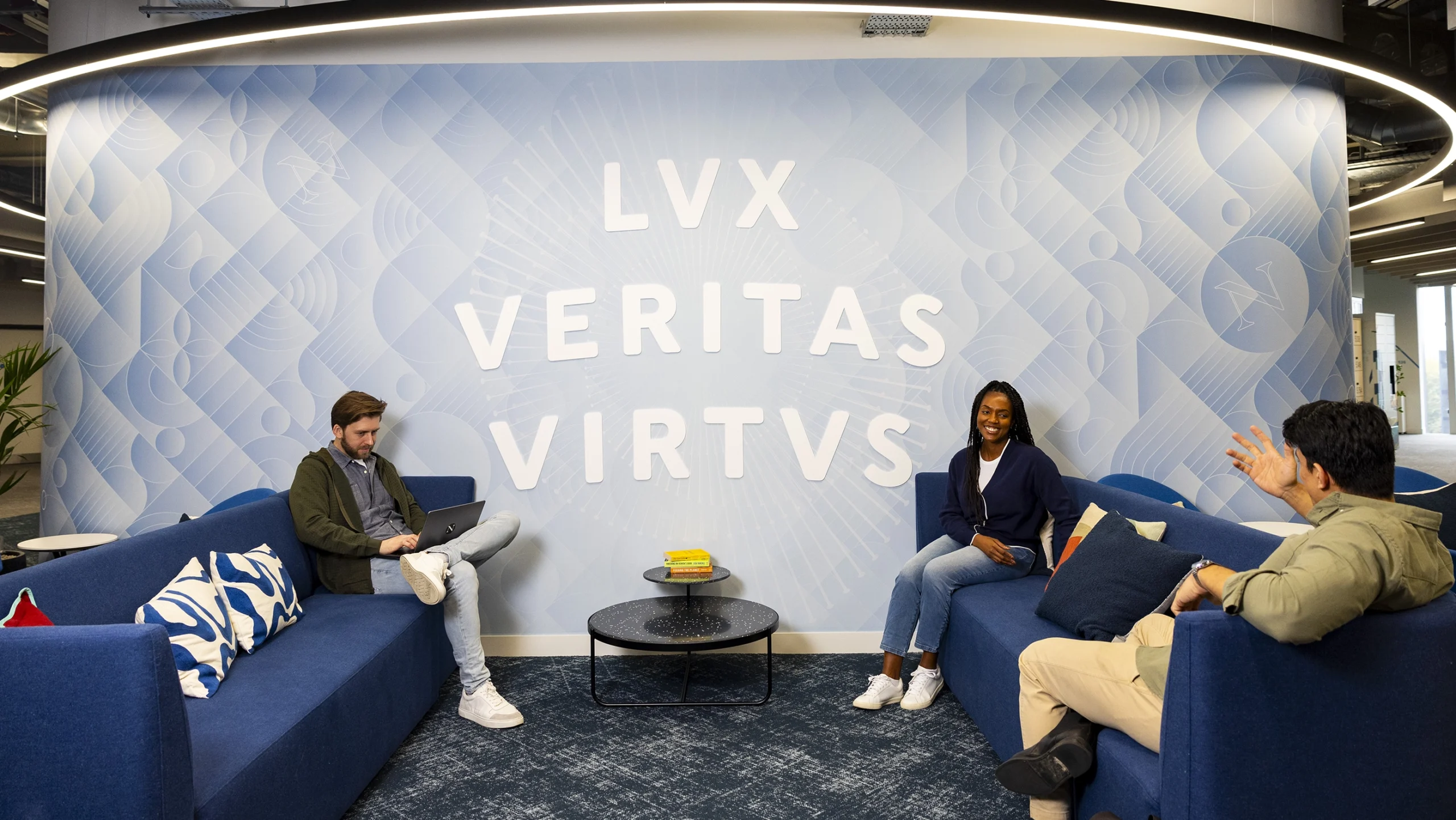 Three people sit on blue couches in a modern lounge area with a round coffee table, discussing Master’s Tuition Fees. The wall behind them reads "LVX VERITAS VIRTUS" in large white letters on a light blue geometric background.