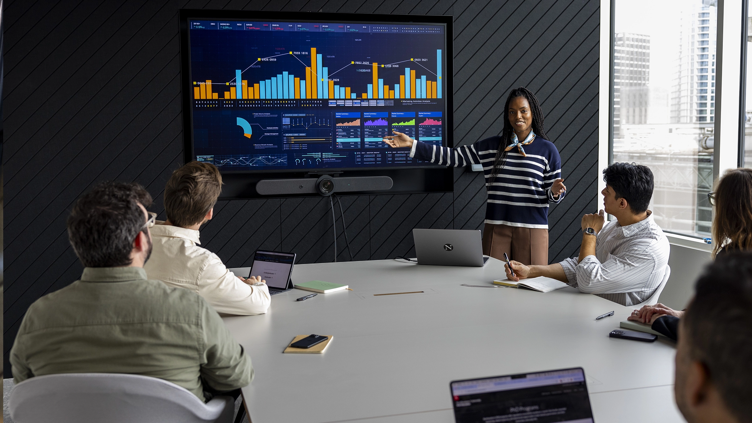 A woman with expertise in masters-degrees stands and presents colorful data charts on a large screen to four seated colleagues in a modern conference room with large windows.