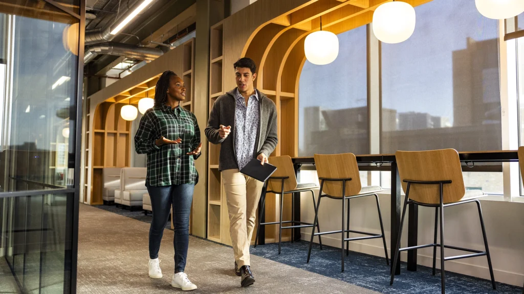 Two people walk and talk in a modern office corridor with large windows, wooden accents, bar stools, and hanging globe lights. The bright, collaborative atmosphere is perfect for discussing ideas or exploring masters-degrees opportunities.