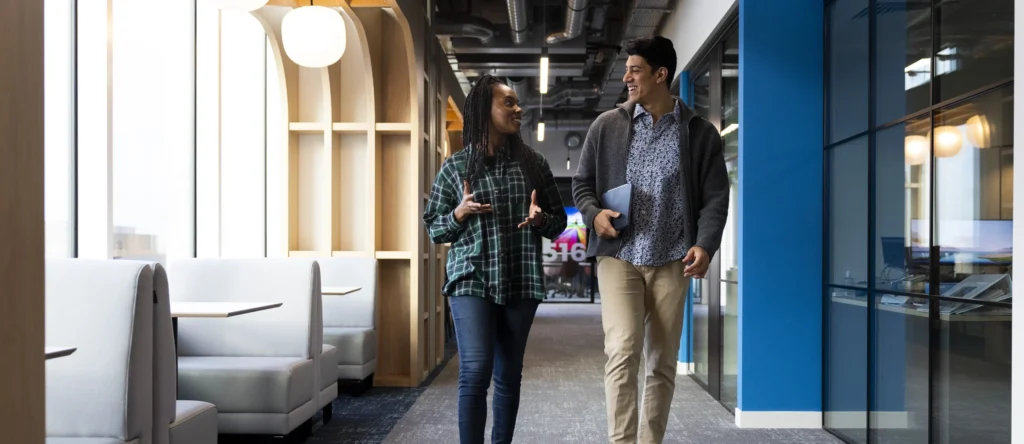 Two people walk together down a modern office hallway, smiling and talking about their masters-degrees, each holding a tablet. The hallway features glass walls, wooden accents, and booth seating on the left side.