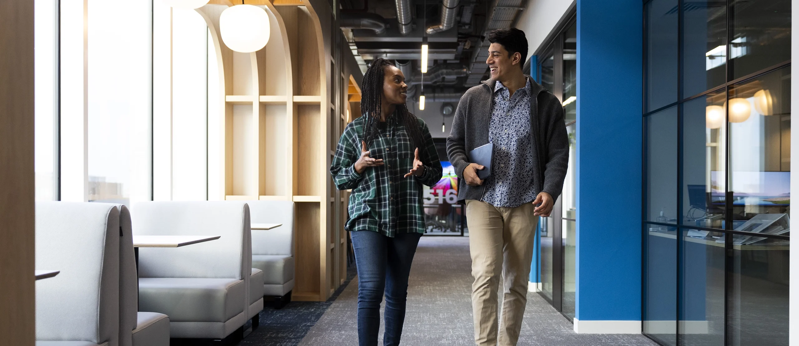 Two people walk together down a modern office hallway, smiling and talking about their masters-degrees, each holding a tablet. The hallway features glass walls, wooden accents, and booth seating on the left side.