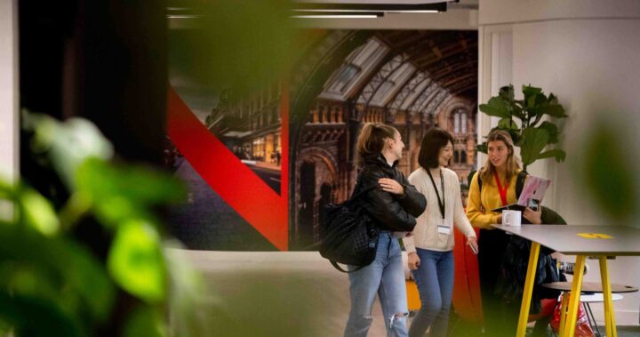 Three young women walk and talk together in a modern indoor space with green plants in the foreground and a colorful mural on the back wall.