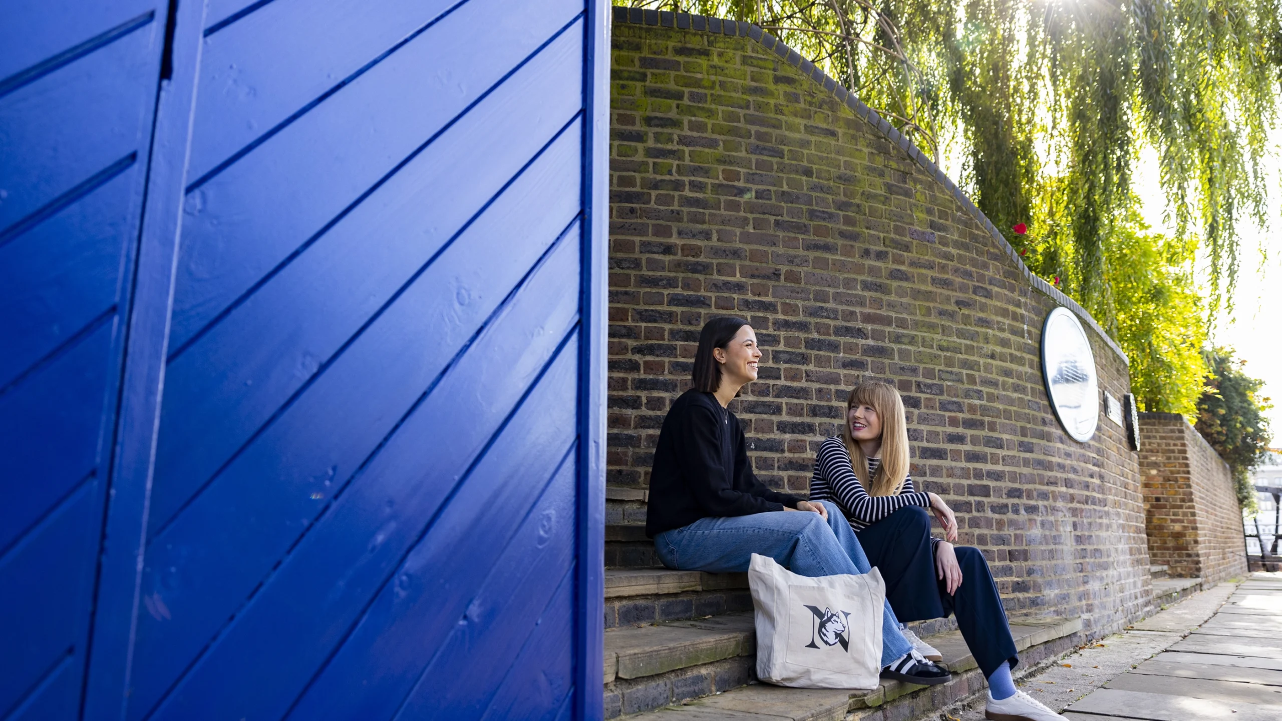 Two women sit on outdoor stone steps by a brick wall, chatting and smiling. Sunlight filters through trees above, and a large blue door occupies the left side of the image. A white tote bag sits near them.
