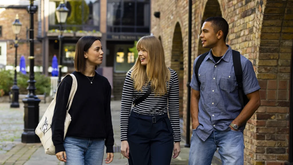 Three young adults, two women and one man, walk together outside along a brick walkway. They are smiling, casually dressed, and each carries a bag. The setting is urban, with brick buildings and street lamps in the background.