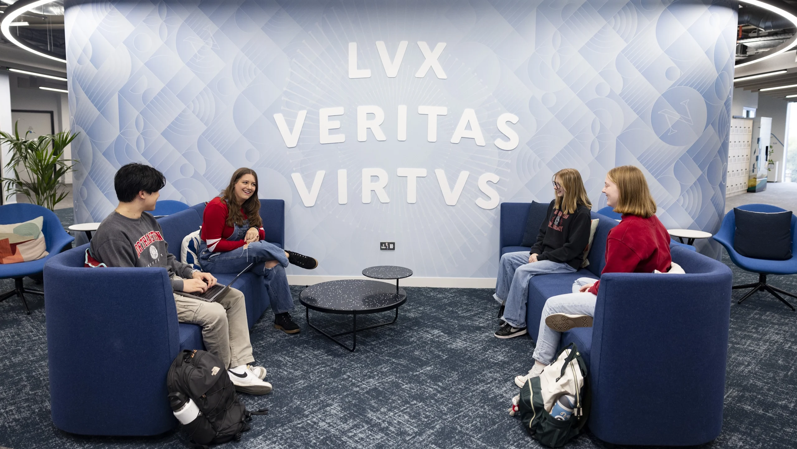Four students sit and talk in blue chairs around small tables in a modern lounge, experiencing campus life beneath a large wall that reads "LVX VERITAS VIRTUS." The space is bright with contemporary design elements.