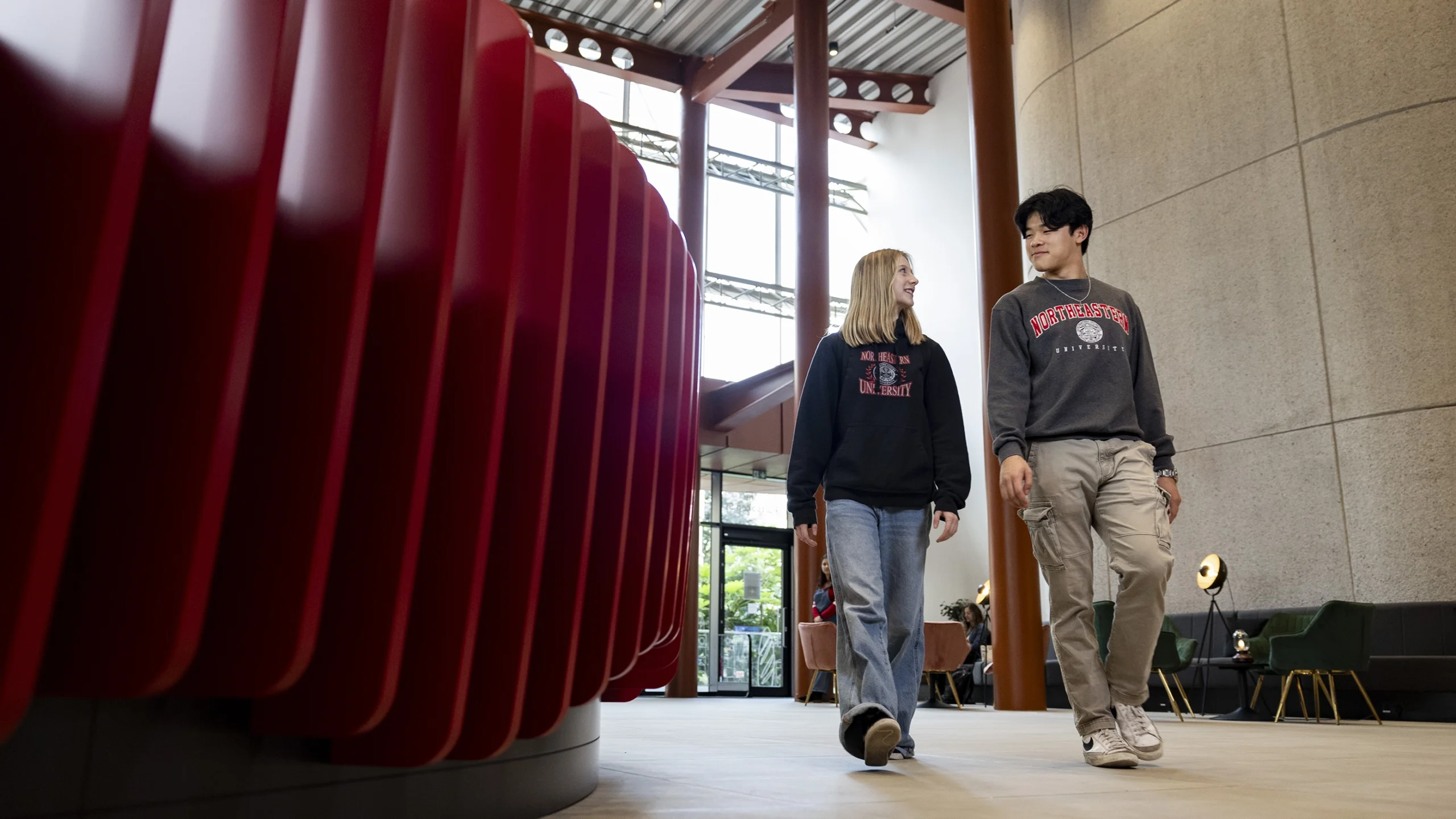Two students wearing Stanford University sweatshirts walk and talk inside a modern building with large windows and red architectural details, discussing their experiences pursuing undergraduate-degrees.