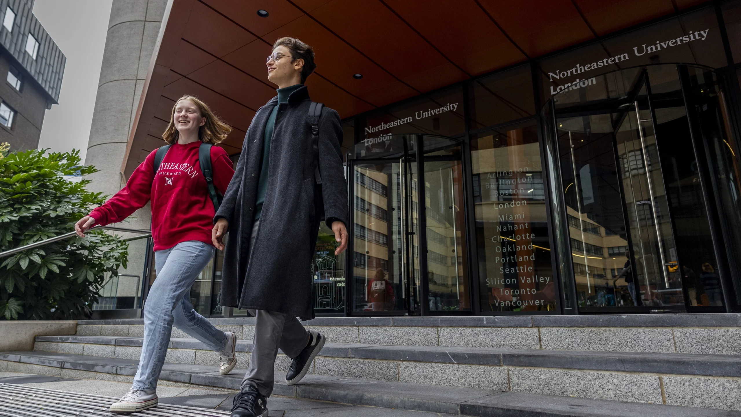 Two students walk down steps outside Northeastern University London. One wears a red sweatshirt and jeans, the other a long coat and scarf. Glass building entrance and university name are visible in the background.