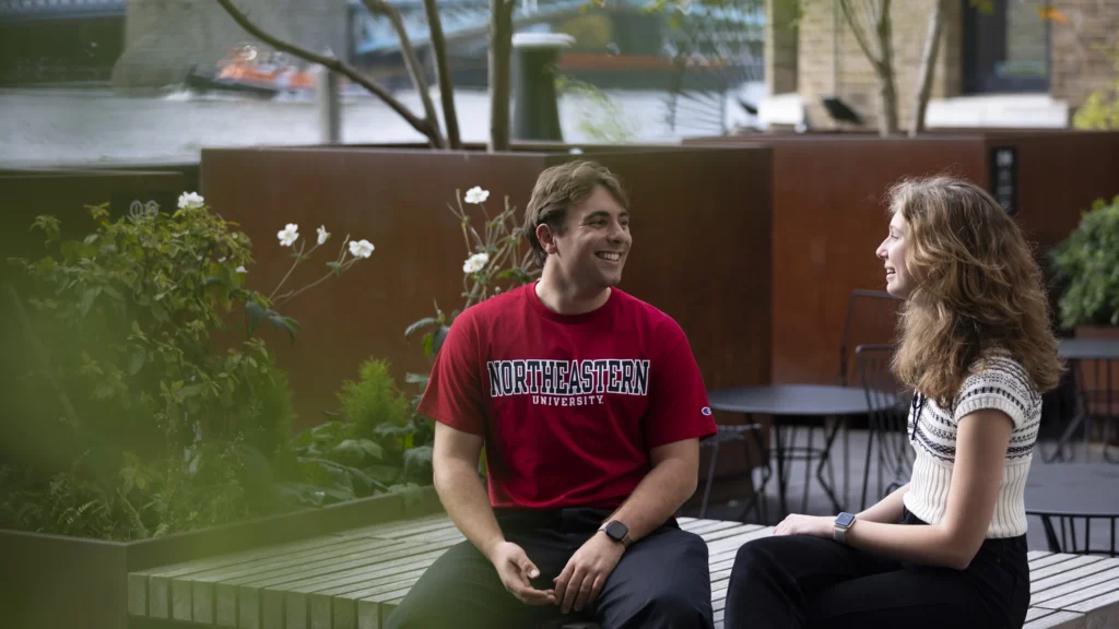 Two young adults sit on outdoor benches, smiling and talking. The man wears a red "Northeastern University" shirt; the woman wears a white and black top. Green plants and a river are visible in the background.