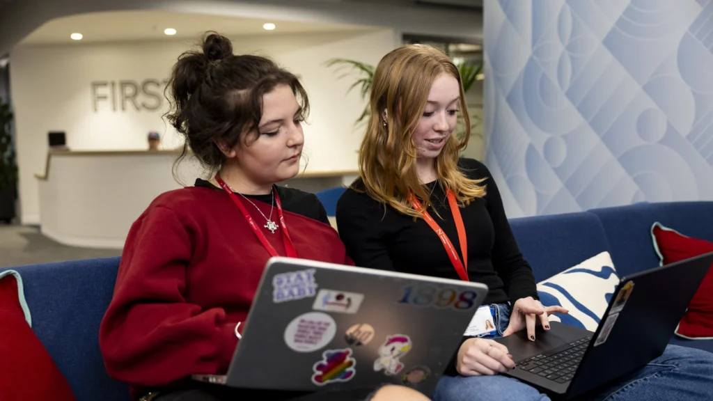 Two young women sit on a blue couch, working together on laptops. One wears a red sweater and the other a black top with a red lanyard. They appear focused and engaged in a collaborative task in a modern office space.