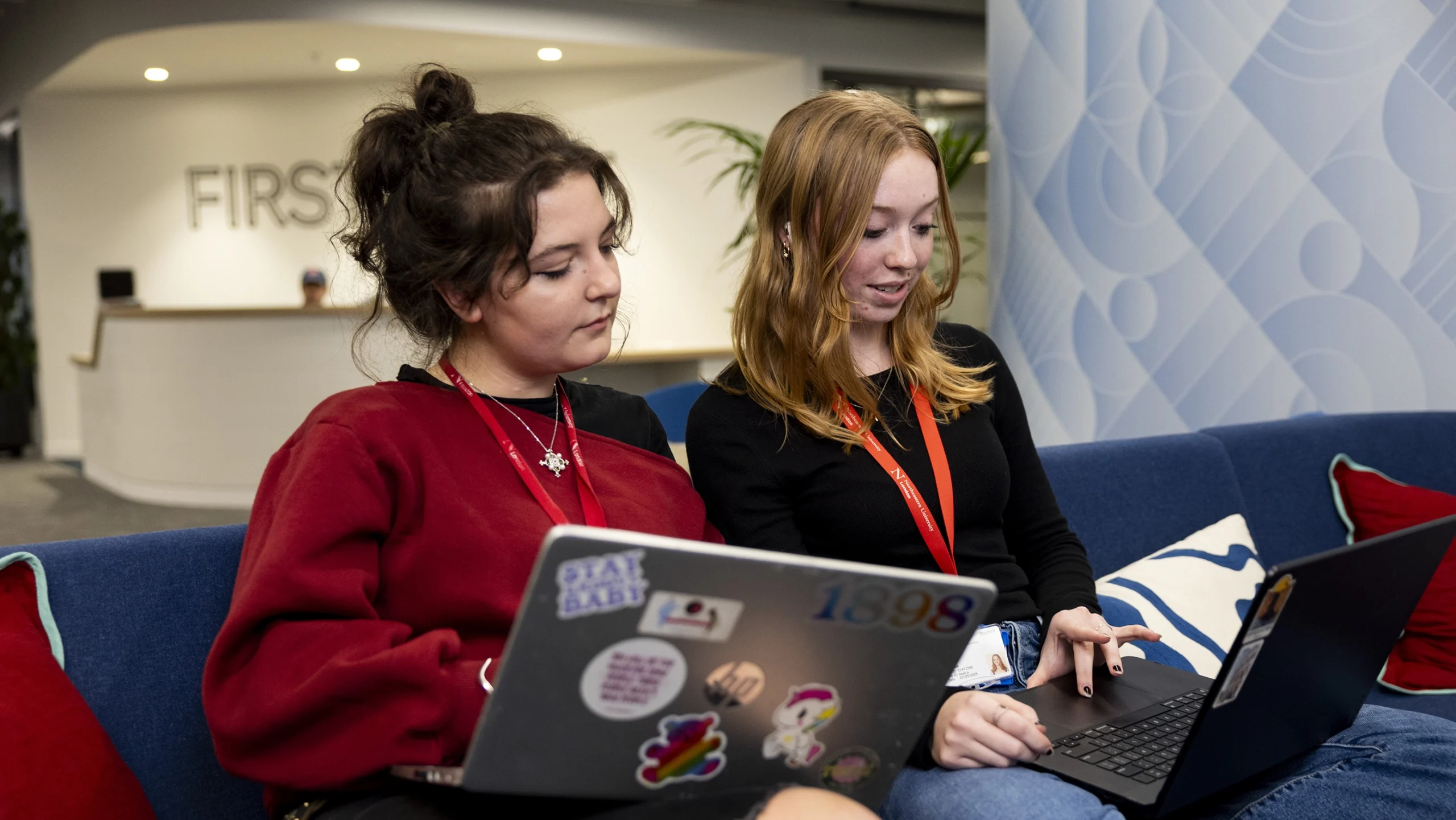 Two young women sit on a blue couch, working together on laptops. One wears a red sweater and the other a black top with a red lanyard. They appear focused and engaged in a collaborative task in a modern office space.