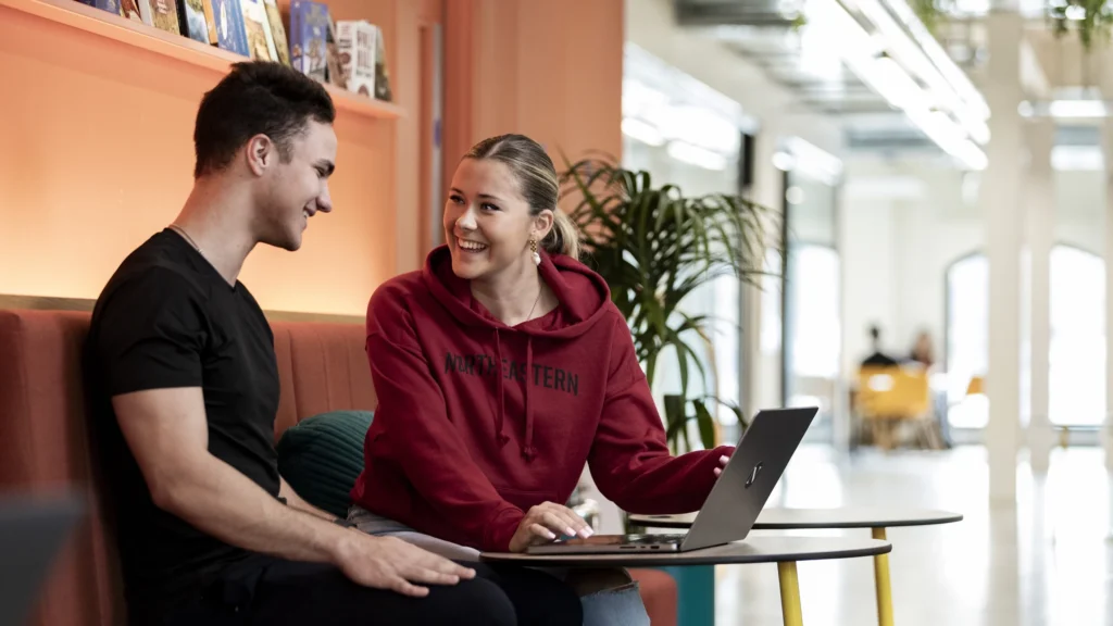 Two young adults sit on a couch in a modern, brightly lit space. The woman, wearing a red “Northeastern” hoodie, is using a laptop and smiling at the man, who is dressed in black. They appear to be having a friendly conversation.