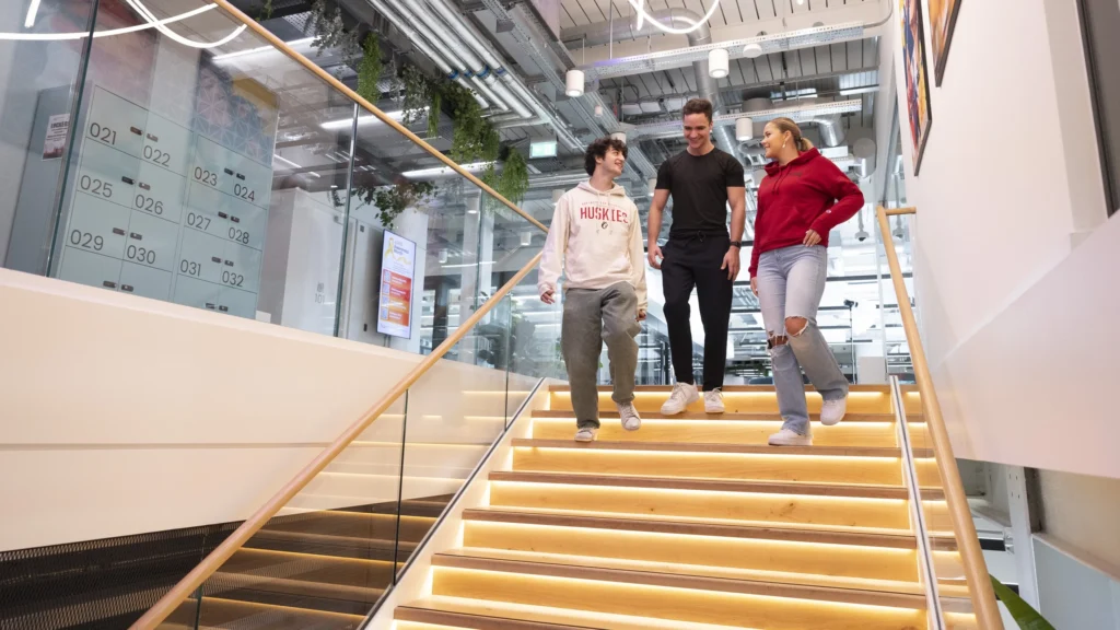 Three people walk down a brightly lit staircase in a modern office building, smiling and talking together. The space has glass walls, plants, and an open, contemporary design.