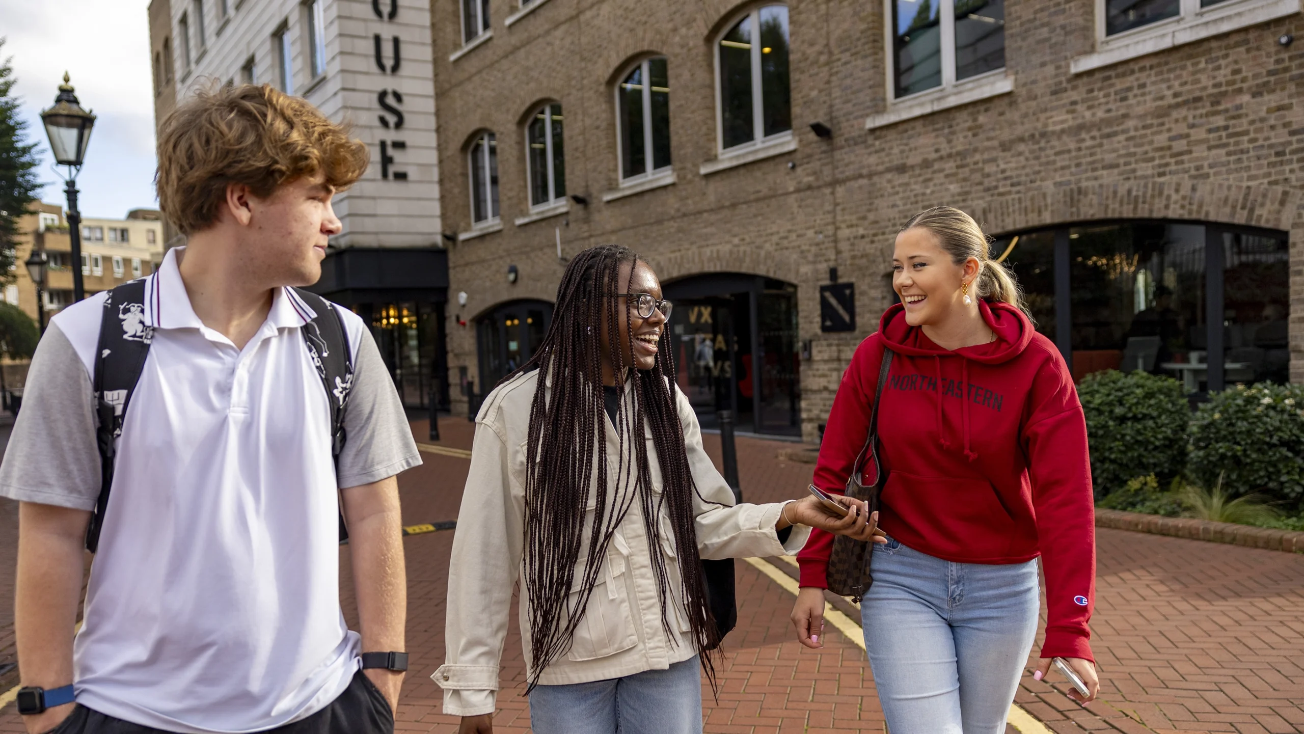 Three college students walk and talk together on a brick sidewalk in front of a building. One wears a white shirt, another a cream jacket, and the third a red "Northeastern" hoodie. All appear happy and engaged.