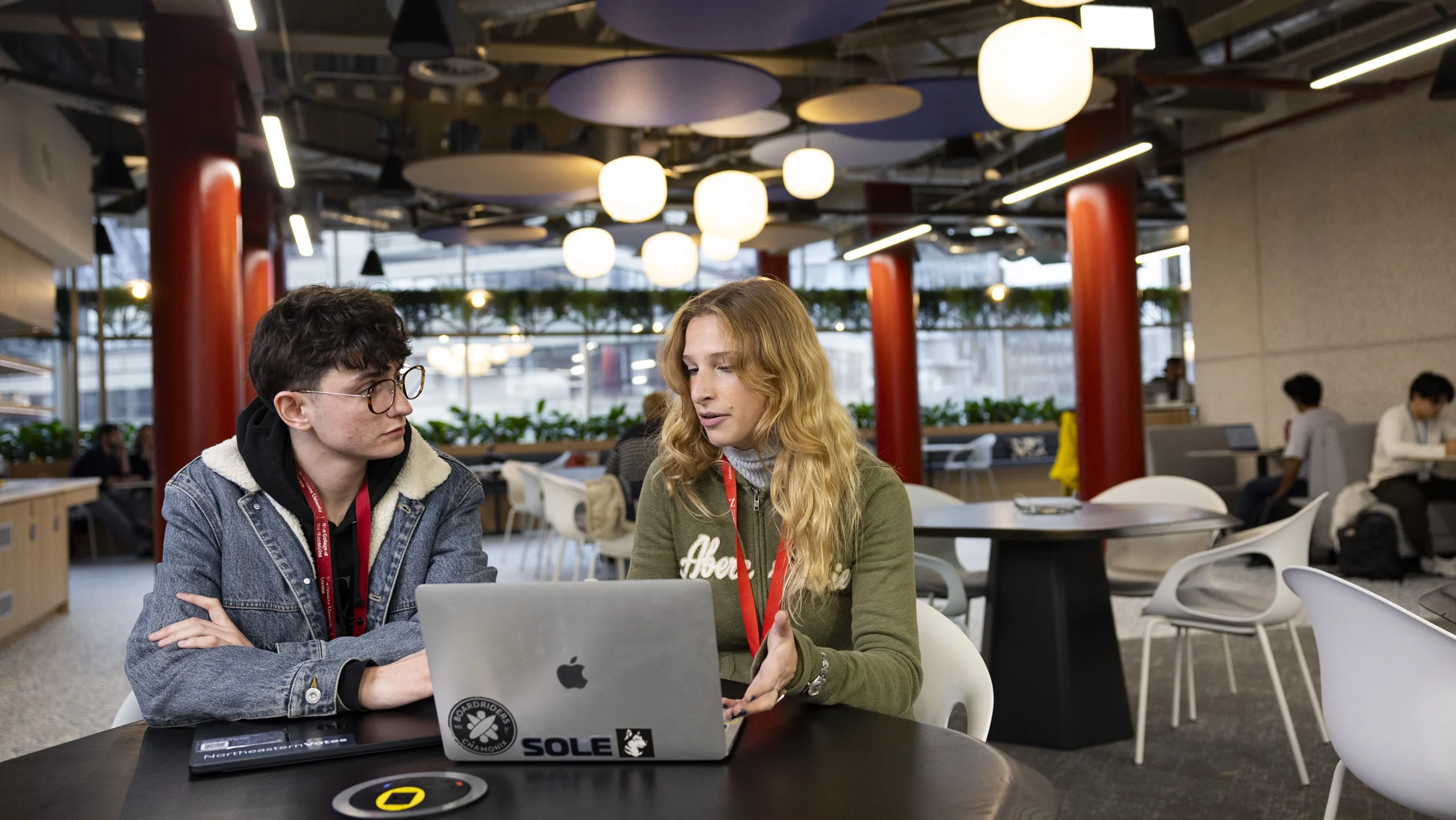 Two students sit at a round table in a modern, brightly lit space, working together on laptops and discussing student money tips. One wears glasses and a jean jacket, the other has long blonde hair and a green jacket. Other people sit in the background.