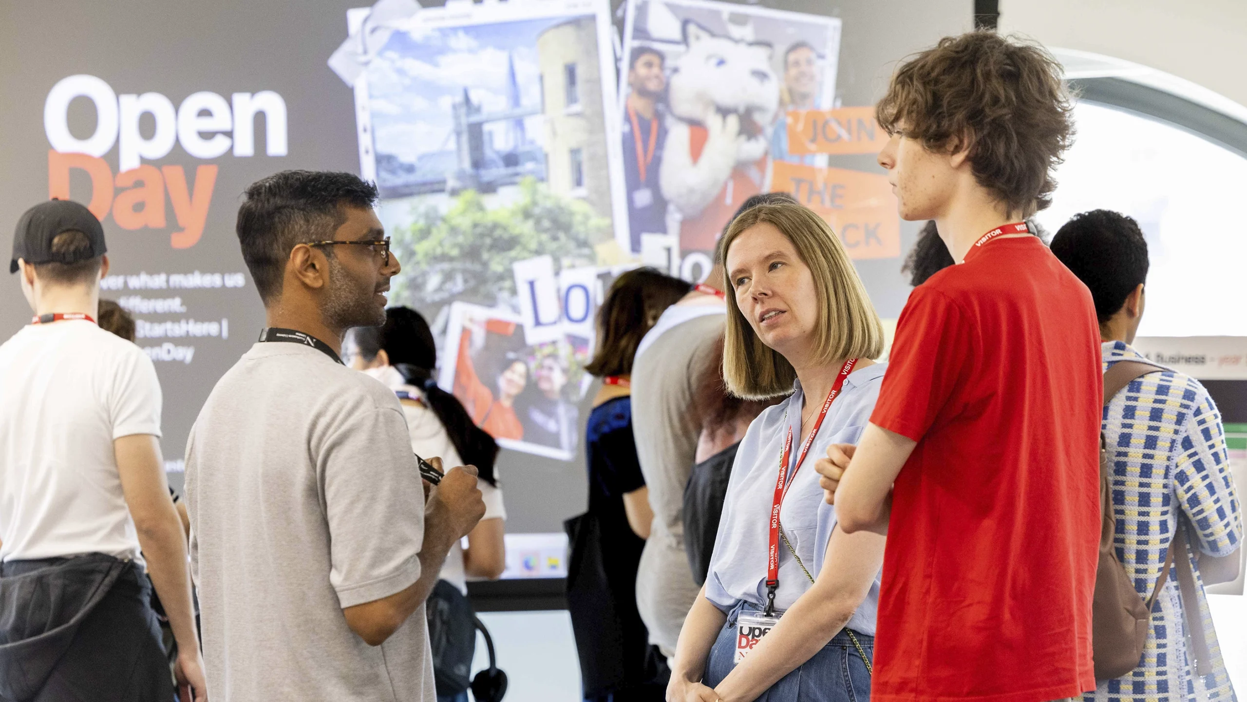 Three people wearing lanyards are talking at an open day event. Other attendees are in the background, with a large "Open Day" sign and collage of photos visible on the wall behind them.