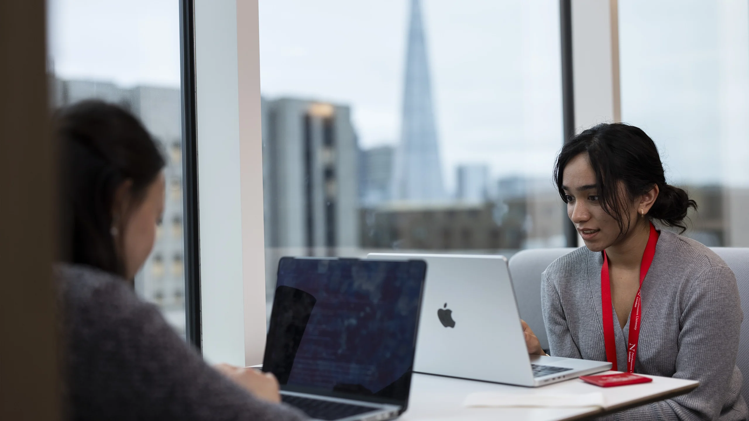 Two women sit at a table in a modern office space, each working on a laptop. Large windows behind them show a cityscape with tall buildings and a pointed skyscraper in the distance.