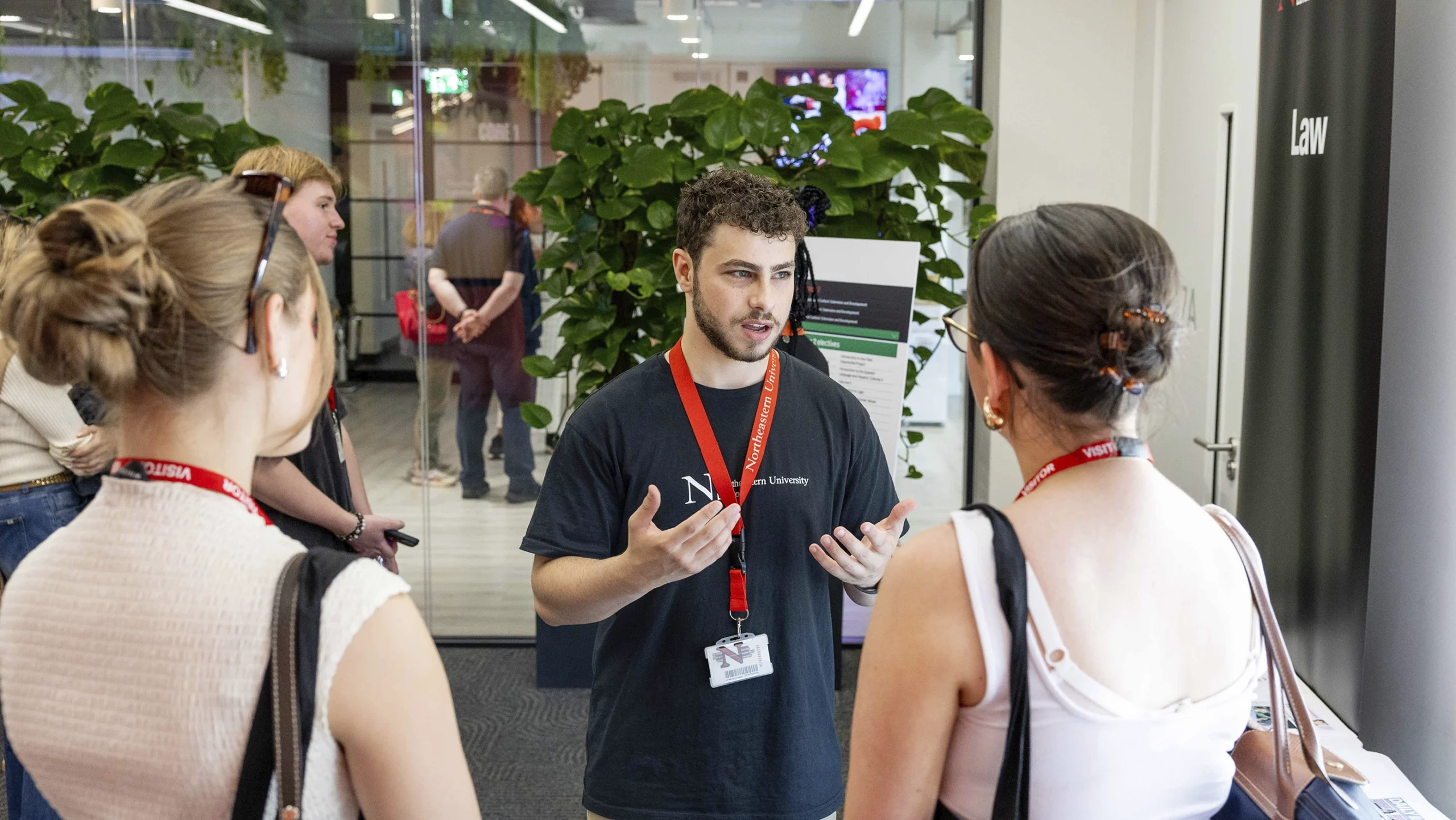 A man with a lanyard speaks to two women wearing lanyards in an indoor setting, with other people and large green plants visible in the background.