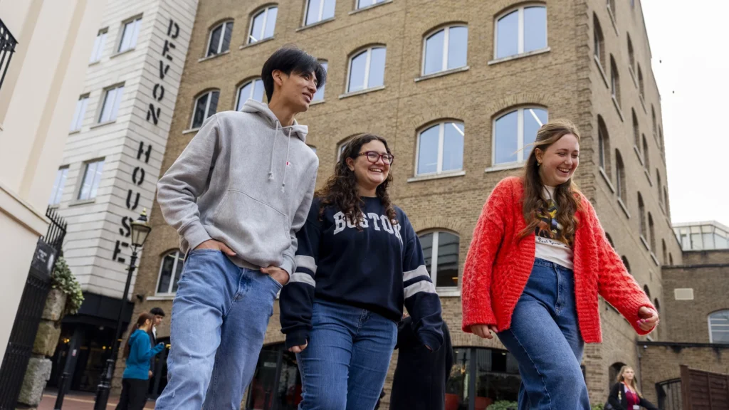 Three young adults walk and smile together outside a brick building with large windows and a sign that reads "Devon House." They appear happy and casually dressed in jeans and sweaters.