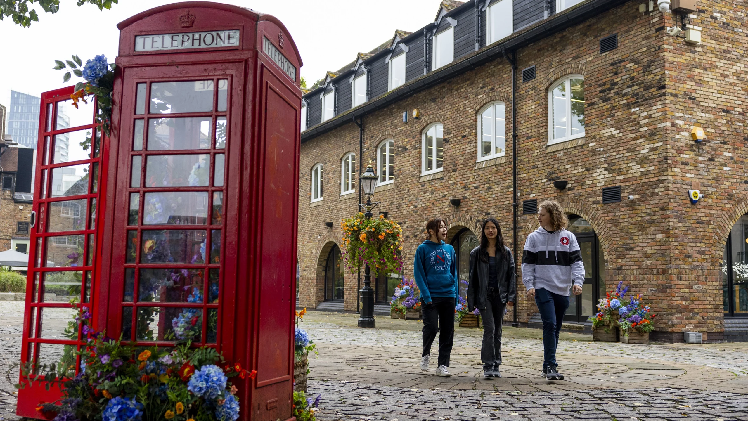 Three people walk on a cobblestone street next to a traditional red British telephone booth decorated with flowers, with a brick building and colorful hanging baskets in the background.