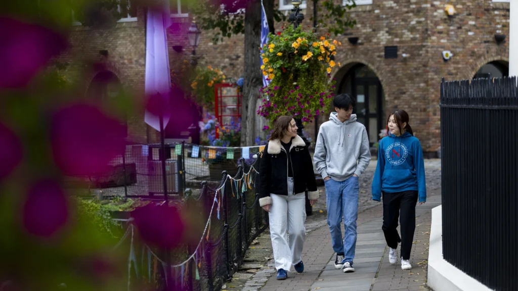 Three young people walk and talk on a path lined with colorful flowers and hanging baskets, next to a brick building, with blurred pink flowers in the foreground.