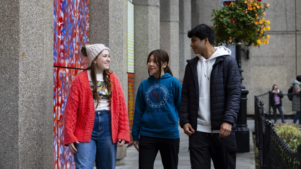 Three young adults walk and smile together on a city sidewalk, passing colorful wall art and a hanging flower basket. The group wears casual, warm clothing. Other people are visible in the background.