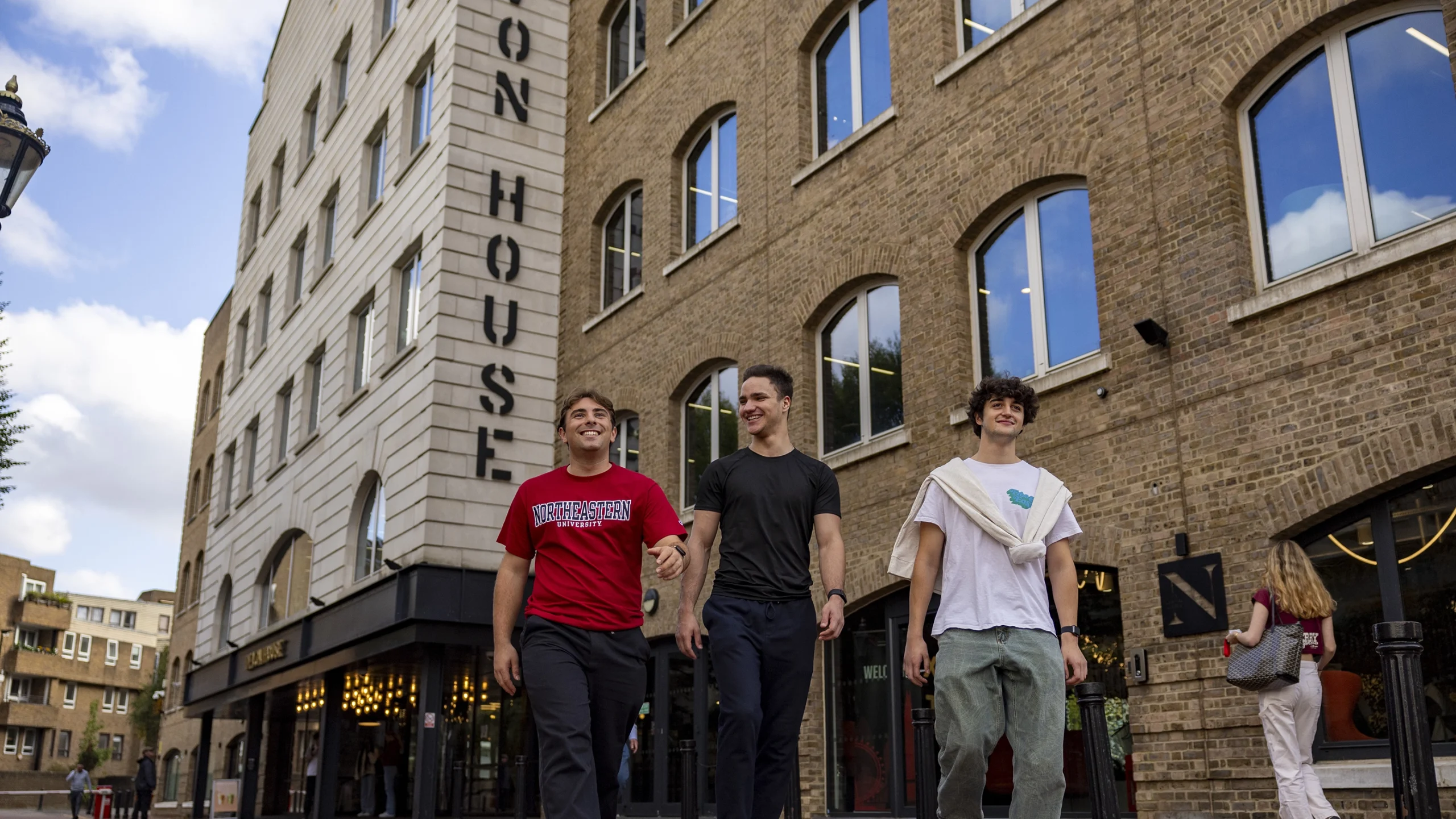 Three young men walk and smile outside a modern brick building with large windows and a sign that reads "ON HOUSE." The sky is clear and blue, and pedestrians are visible in the background.