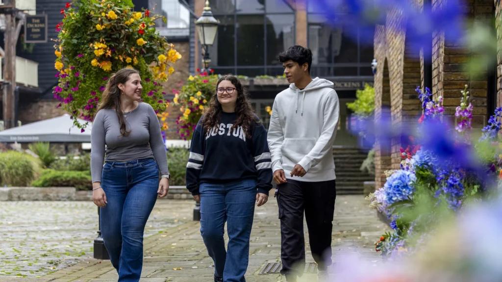 Three young people walk and talk together on a cobblestone path lined with vibrant flowers and hanging baskets, with historic buildings in the background. They appear to be enjoying a pleasant, casual conversation.