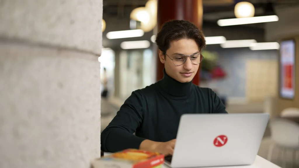 A person wearing glasses and a dark turtleneck sits at a table, working on a laptop with a red logo in a modern, brightly lit office space. Books are visible in the foreground.