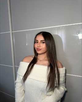 A young woman with long, straight brown hair wears an off-shoulder white sweater, standing indoors against a gray wall with white grid lines. She is looking at the camera with a slight smile.
