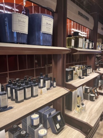 Shelves display various bottles, jars, and boxes of perfumes and candles in a store, with items neatly arranged and labeled. During a student visit to Atelier Rebul, the warm wooden shelves and inviting ambiance create a memorable experience.