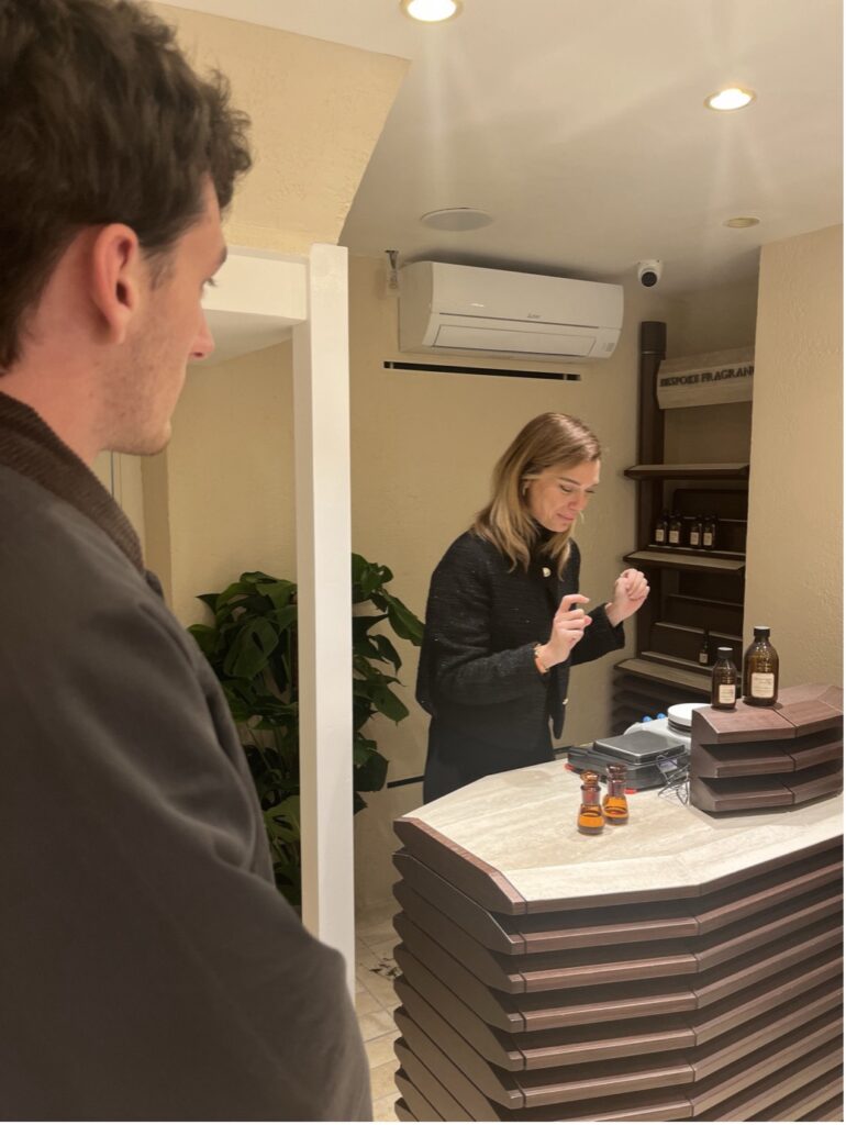 During a student visit to Atelier Rebul, a woman stands behind a curved wooden counter with bottles on it, holding a tester strip. A man in a dark jacket stands in the foreground, observing her in the boutique setting.
