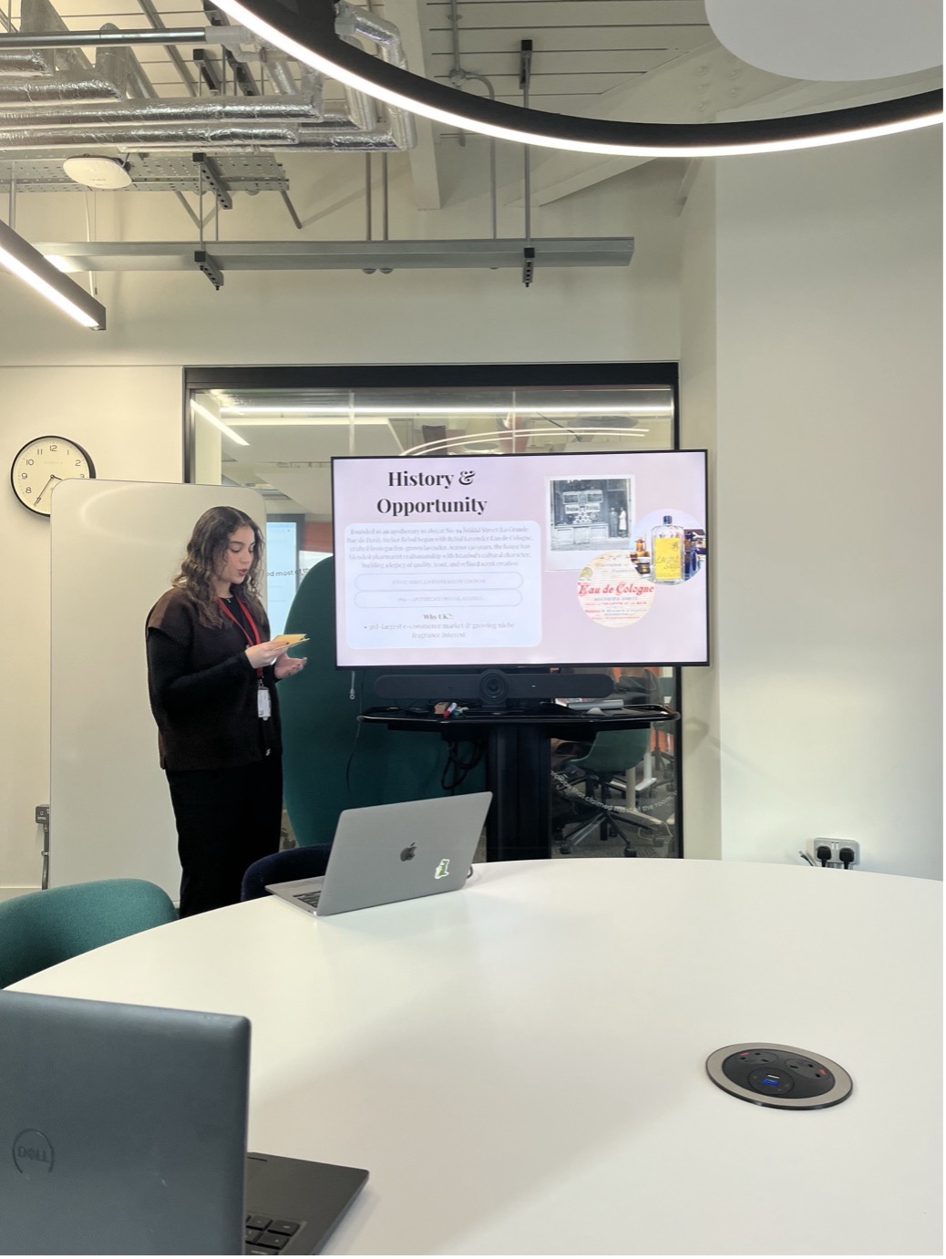 A woman stands by a screen giving a presentation titled "History & Opportunity" during a Student Visit to Atelier Rebul in a modern conference room with a round table, laptops, and a visible wall clock.