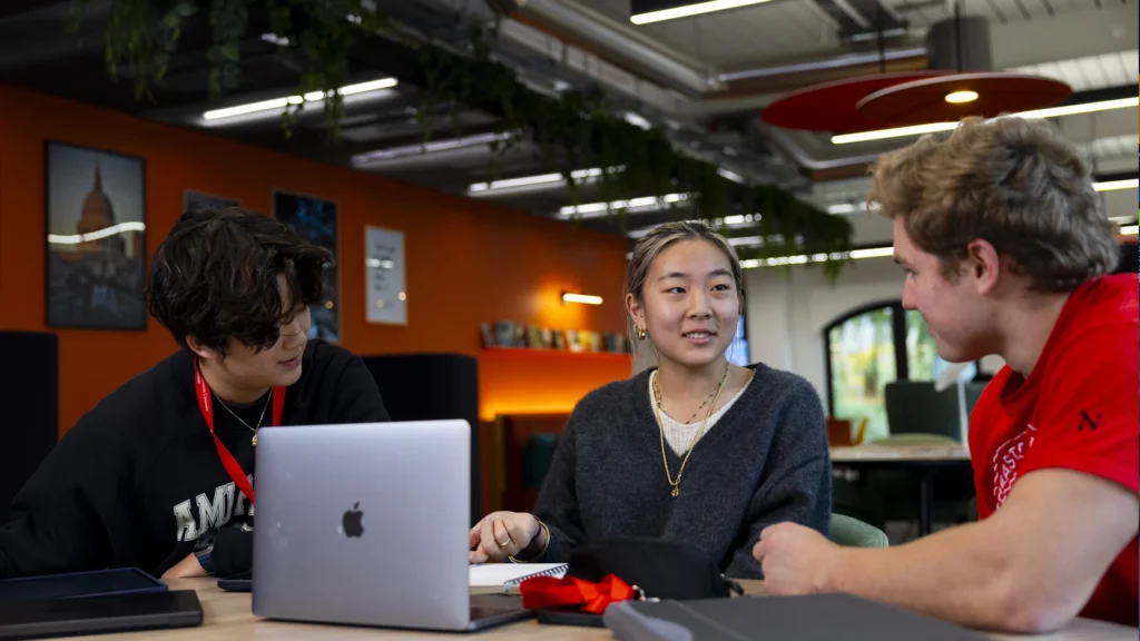 Three young adults sit around a table in a modern, brightly-lit room, talking and smiling. An open laptop, notebooks, and pens are on the table, suggesting a study or collaborative work session.