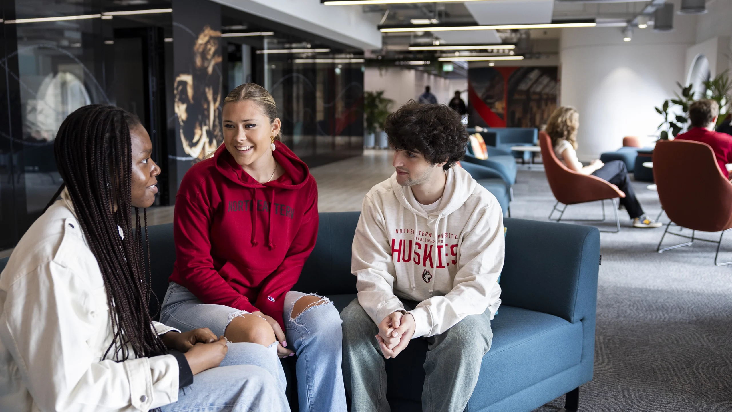 Three students sit on a blue couch in a modern lounge, talking and smiling. Other people are seated in the background. The atmosphere appears friendly and relaxed.