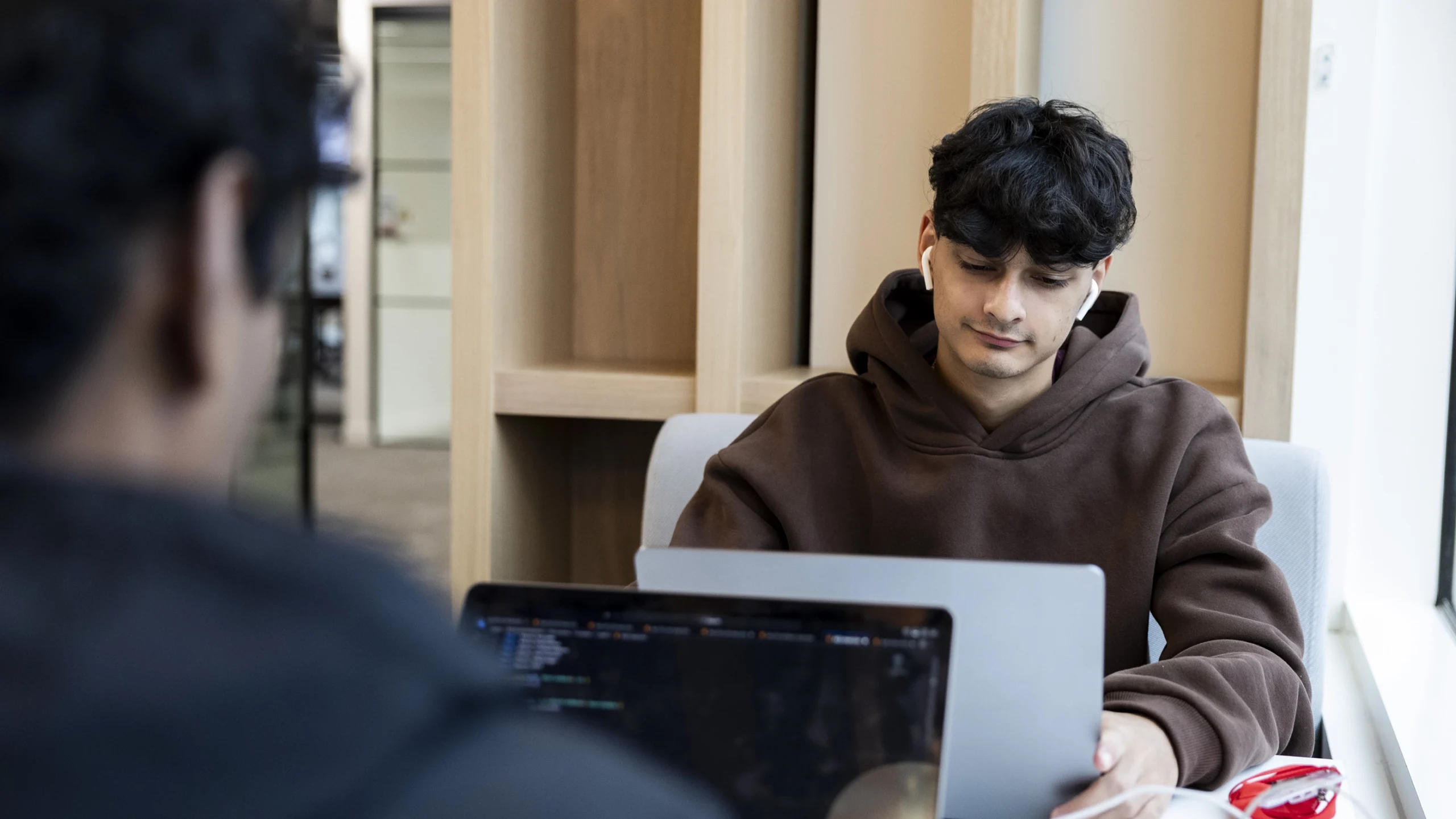 A young person in a brown hoodie sits at a table, focused on their laptop. Another person is partially visible in the foreground, also using a laptop. The setting appears to be a modern, indoor workspace.