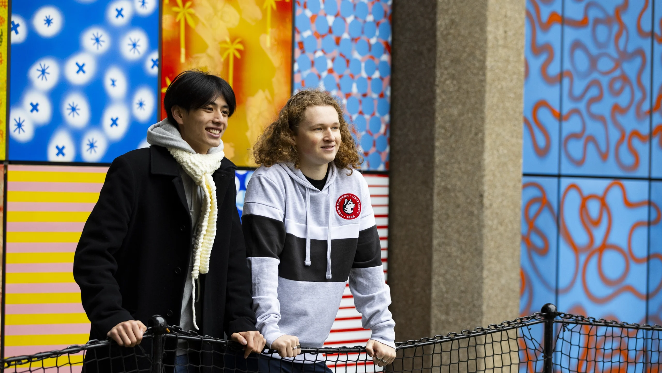 Two people stand outside, smiling and leaning on a black fence. Colorful, patterned tiles decorate the wall behind them. One person wears a scarf and coat, the other a hoodie with stripes.