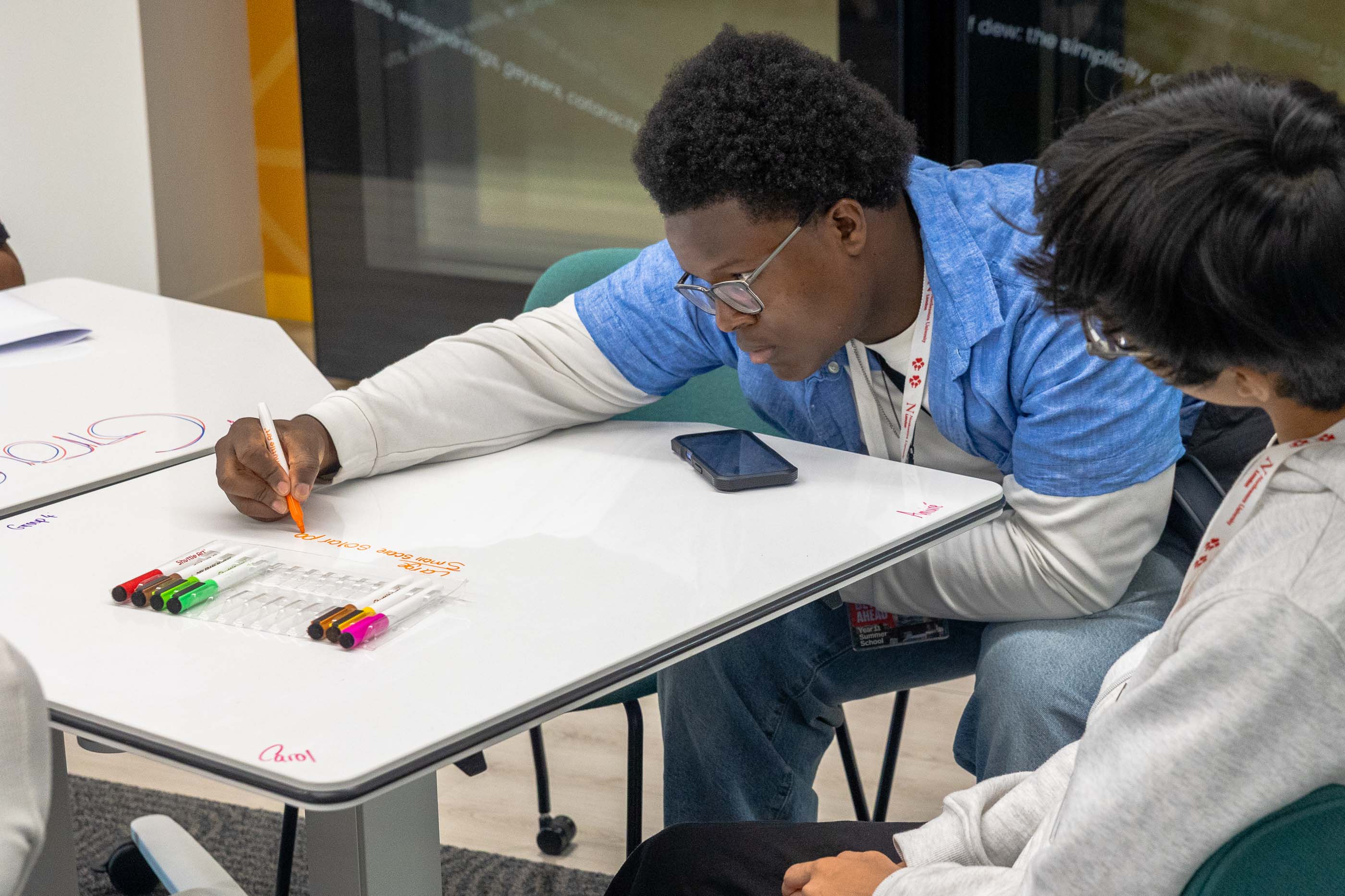 summer_school_2025-1-1 A student at summer school writes on a whiteboard table with markers, focused on his work, while another student sits nearby. A smartphone lies on the table as they collaborate in a classroom or study setting.