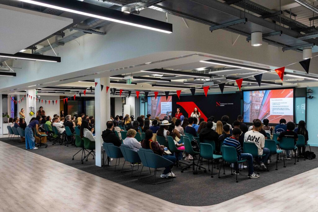 A large group of people sit in a modern, open space for a Summer School session, facing a screen and speaker giving a presentation. Red and black bunting decorates the area. The audience appears engaged and attentive.