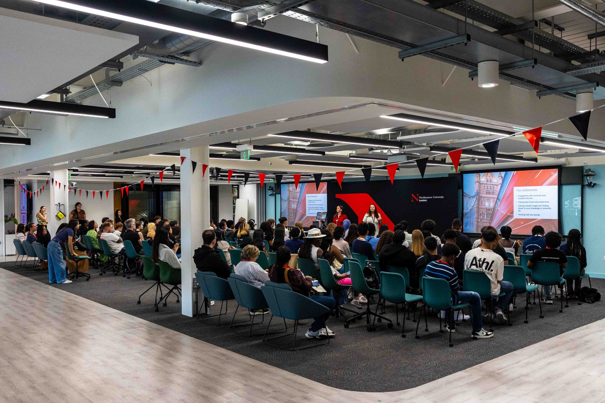 A large group of people sit in a modern, open space for a Summer School session, facing a screen and speaker giving a presentation. Red and black bunting decorates the area. The audience appears engaged and attentive.