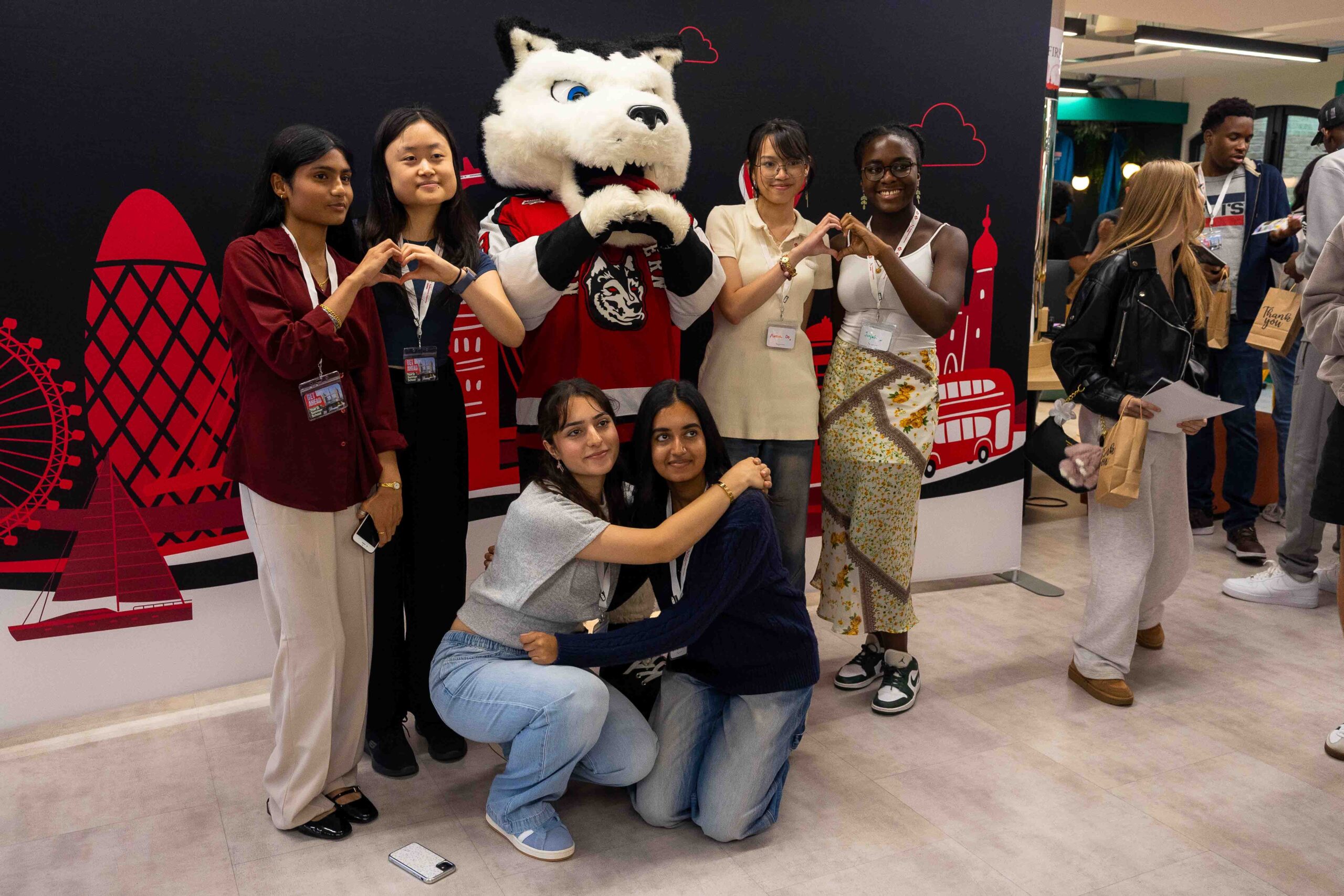 summer_school_2025-18 A group of six Summer School students pose with a husky mascot, making heart shapes with their hands. Two students kneel in front, hugging. The background features red illustrations of London landmarks in the lively space.