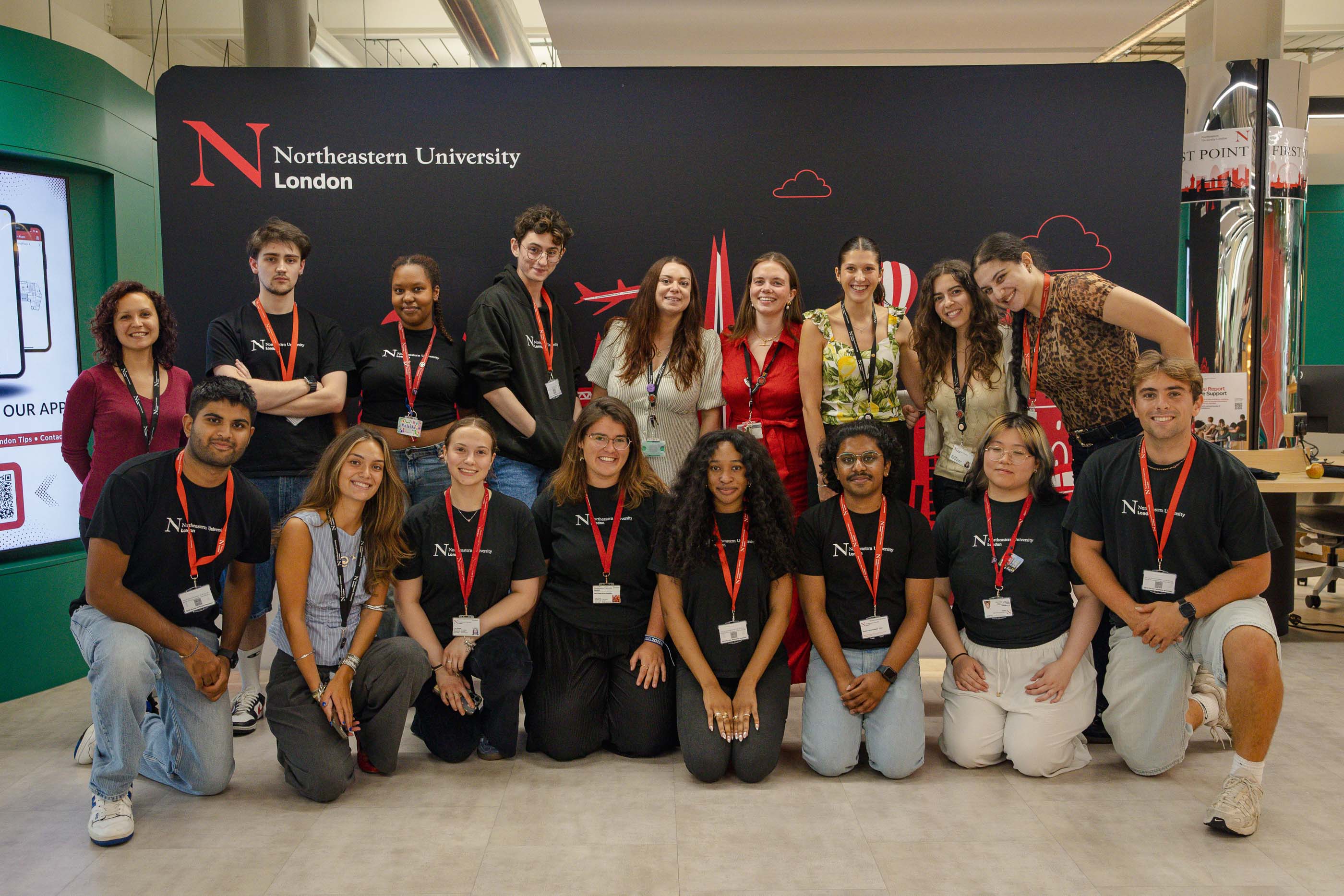 summer_school_2025-20-1 A group of sixteen diverse young adults and two older women, all wearing name badges, pose and smile together in front of a Northeastern University London Summer School backdrop, some standing and some kneeling.