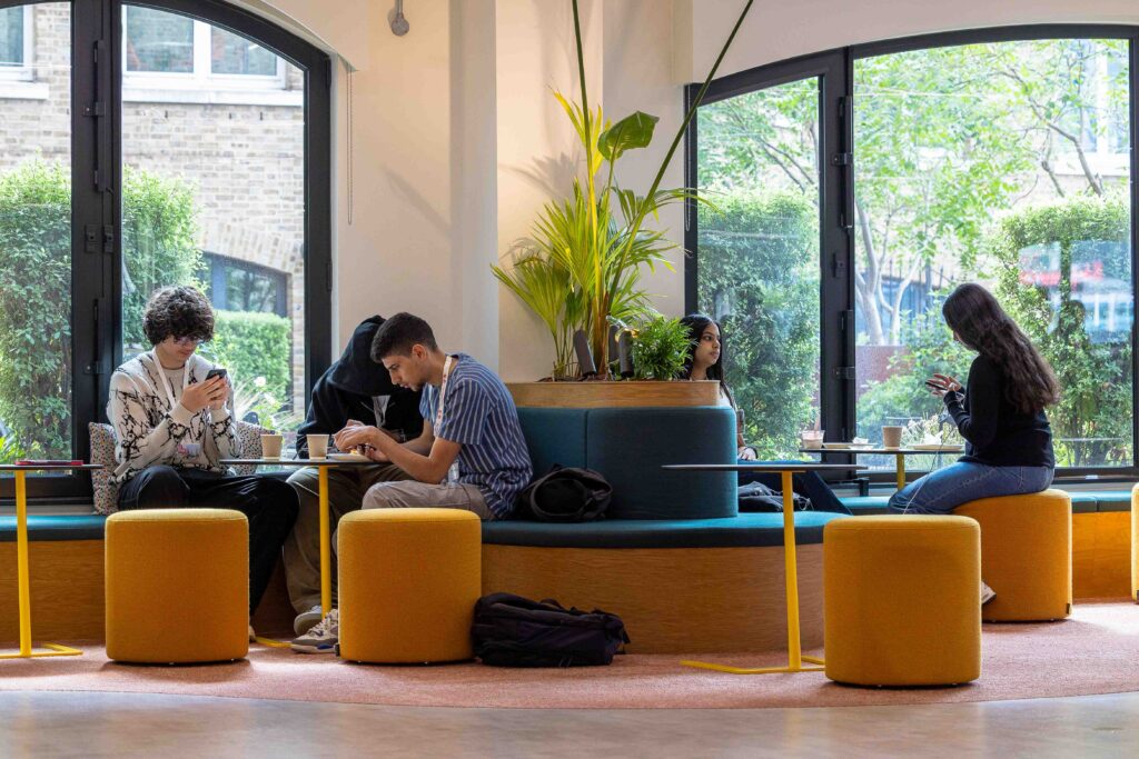 Four people sit on a curved bench in a modern, brightly lit cafe, each focused on their phone or laptop—perhaps catching up on Summer School assignments. Large windows reveal greenery outside, and yellow stools surround the tables.