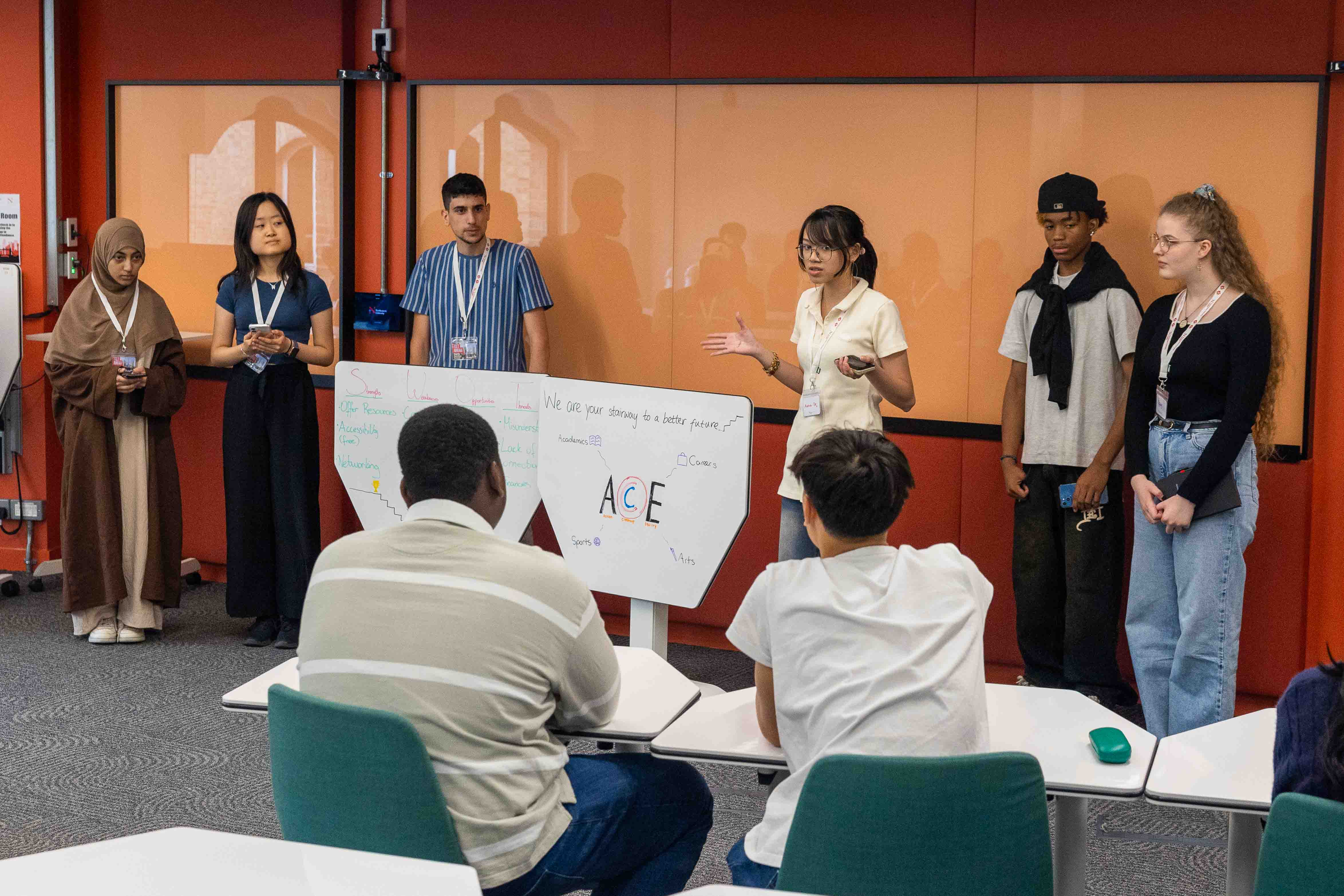 summer_school_2025-9 A group of six young people stand and present in front of a whiteboard with writing, while three others sit facing them in a Summer School classroom with orange walls.
